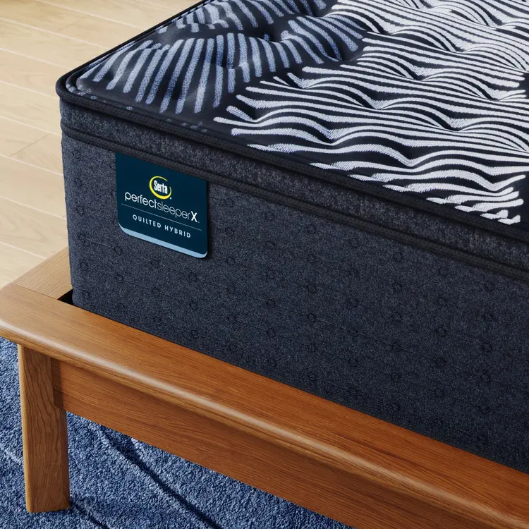 Corner of Perfect Sleeper X Quilted Hybrid Max Firm Pillow Top Mattress sitting in a brown bedframe || feel: Firm Pillow Top || level: Level 2