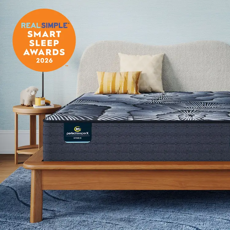 A Perfect Sleeper X Hybrid mattress on a wooden bed frame in a styled bedroom scene with a side table, pillows, rug, and a light blue wall. An orange badge in the top left reads 'REAL SIMPLE SMART SLEEP AWARDS 2026.