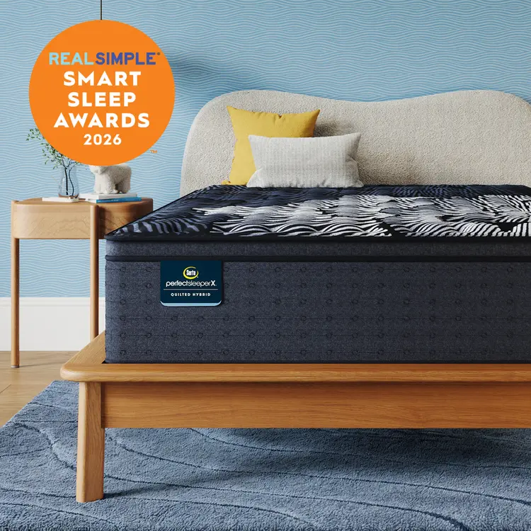 A Queen-sized Perfect Sleeper X Quilted Hybrid mattress with a dark navy and white patterned top surface is shown on a wooden bed frame in a styled bedroom. The background wall is textured light blue, with a beige upholstered headboard, two accent pillows (one yellow, one white), a matching blue rug, and a small wooden side table.